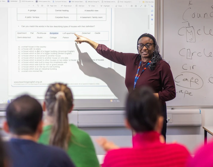 A female lecturer wearing glasses and a lanyard points to a projected lesson on a whiteboard while teaching a group of students. A female lecturer wearing glasses and a lanyard points to a projected lesson on a whiteboard while teaching a group of students.
